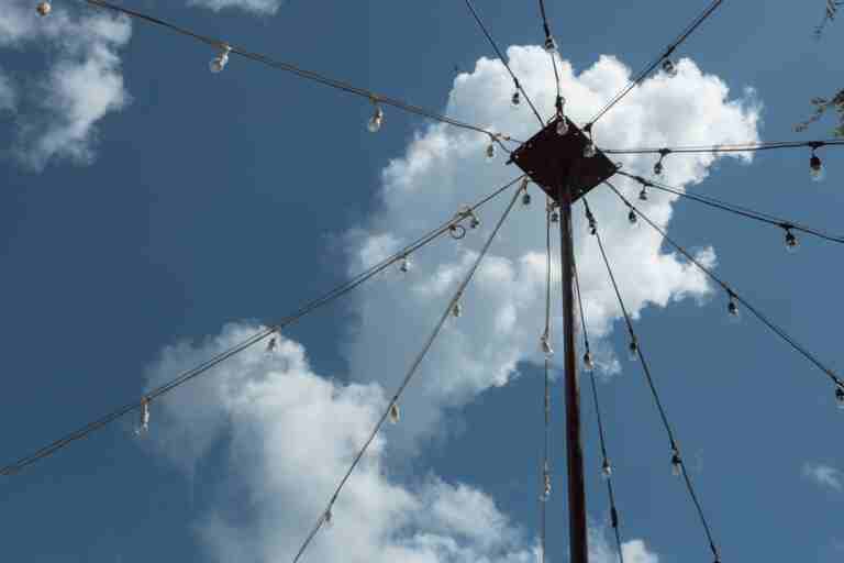 Low angle view of string lights on a post with fluffy clouds and a clear blue sky background.